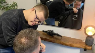 Mokinys kirpykloje trimeriu tvarko kliento barzdą